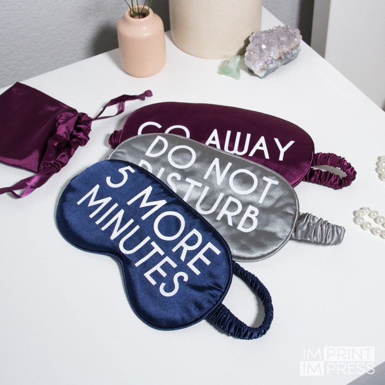 May include: Three sleep masks with different sayings on them. The first mask is a dark purple color with the text 'Go Away' in white. The second mask is a light gray color with the text 'Do Not Disturb' in white. The third mask is a dark blue color with the text '5 More Minutes' in white.