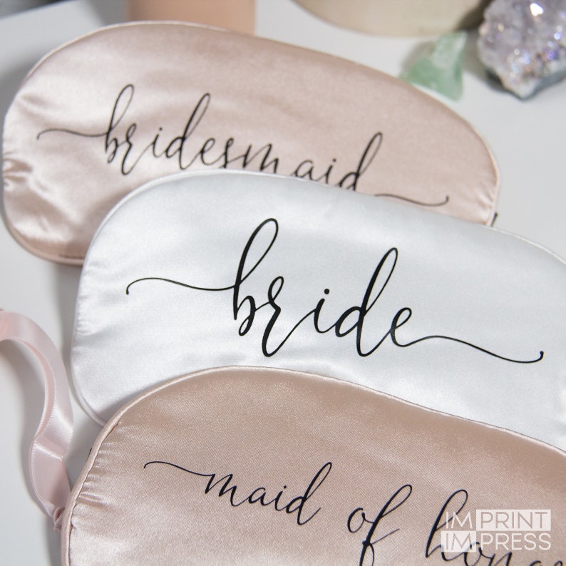 May include: Three satin sleep masks with the words "bridesmaid", "bride", and "maid of honor" printed in black script.