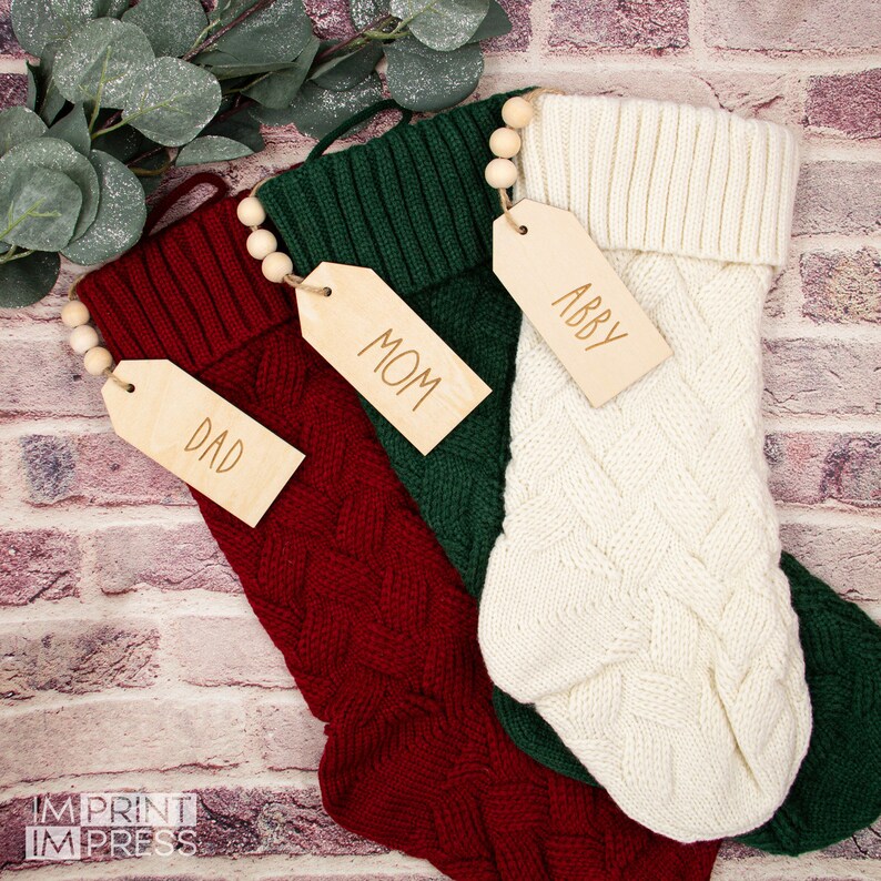 May include: Three knitted Christmas stockings with wooden tags. The stockings are red, green, and white. The tags are labeled "Dad", "Mom", and "Abby".