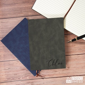May include: A dark gray leather-bound journal with the name "Chloe" embossed in black on the front cover. The journal is lying on a wooden surface with a blue journal and a pen.