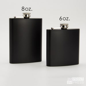 May include: Two black metal hip flasks with silver caps and hinges. The flask on the left is labeled "8 oz." and the flask on the right is labeled "6 oz."