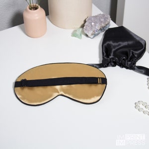 Op de afbeelding: Een gouden satijnen slaapmasker met een verstelbare zwarte elastische band. Het masker ligt op een wit oppervlak.