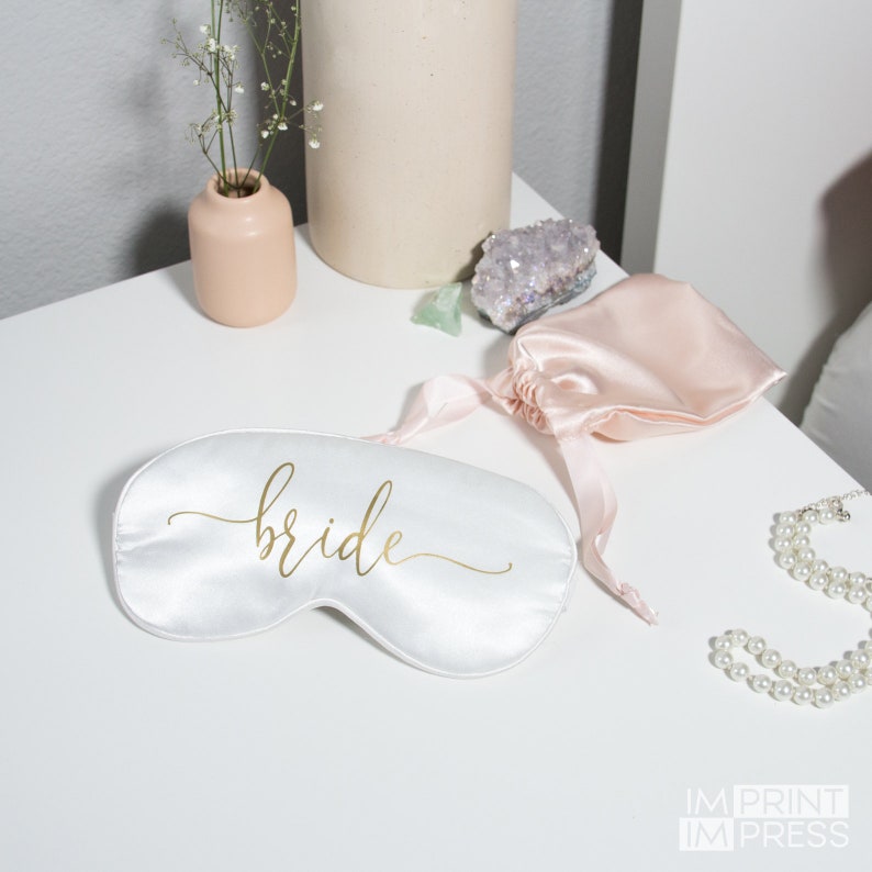 May include: White satin sleep mask with the word "bride" written in gold script. The mask is lying on a white surface with a pink satin bag and a pearl necklace.