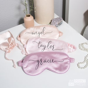 May include: Three pink satin sleep masks with black script lettering. The masks are labeled "angel", "taylor", and "gracie".