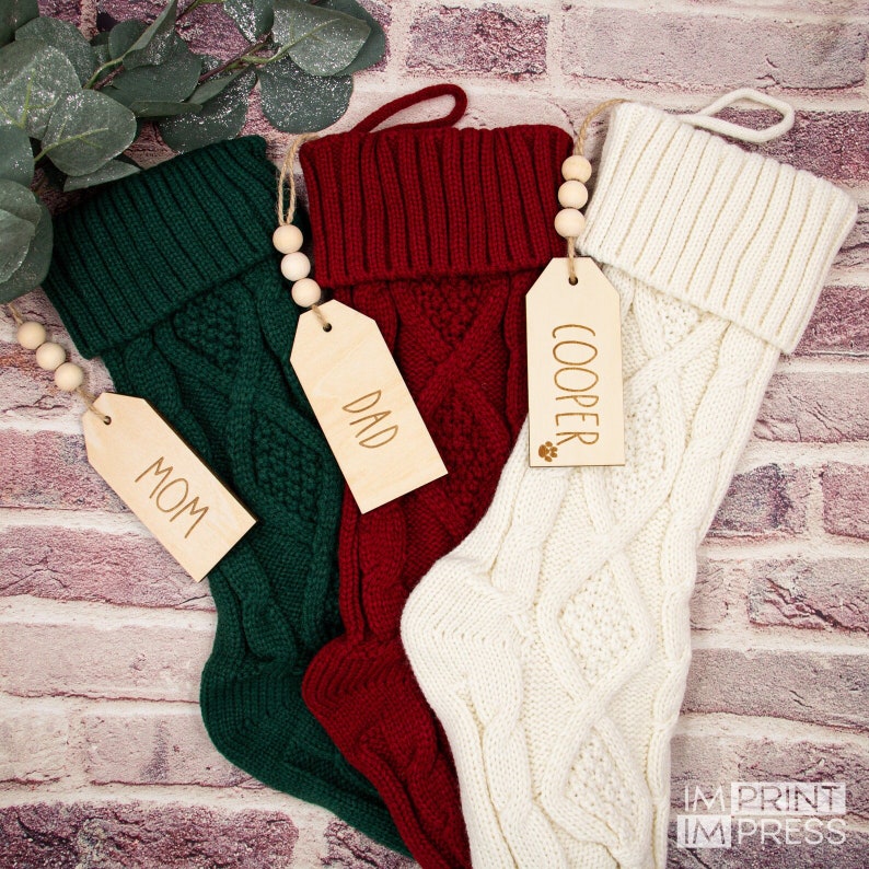 May include: Three knitted Christmas stockings with personalised wooden tags. The stockings are green, red, and white. The tags read "Mom", "Dad", and "Cooper".