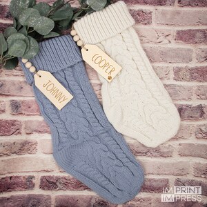 K&ouml;nnte beinhalten: Zwei gestrickte Weihnachtsstr&uuml;mpfe mit Holzetiketten. Der Strumpf links ist blau und hat ein Etikett mit der Aufschrift "JOHNNY". Der Strumpf rechts ist wei&szlig; und hat ein Etikett mit der Aufschrift "COOPER".