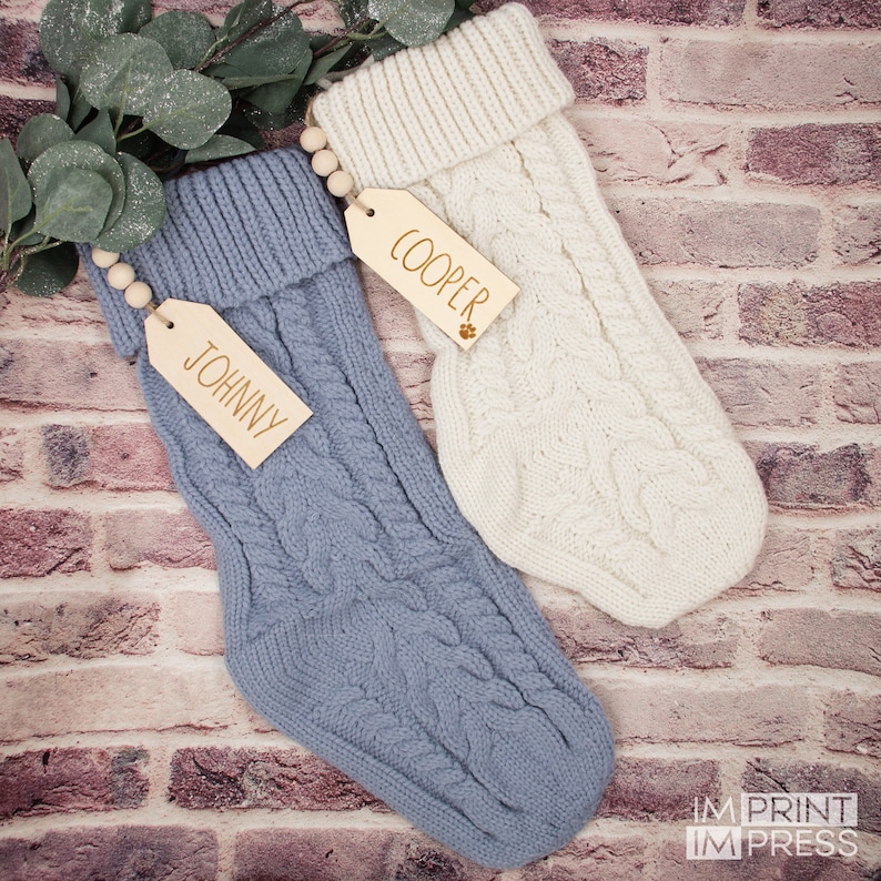 K&ouml;nnte beinhalten: Zwei gestrickte Weihnachtsstr&uuml;mpfe mit Holzetiketten. Der Strumpf links ist blau und hat ein Etikett mit der Aufschrift "JOHNNY". Der Strumpf rechts ist wei&szlig; und hat ein Etikett mit der Aufschrift "COOPER".