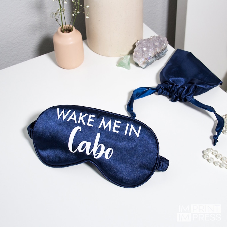 May include: A navy blue satin sleep mask with white text that reads "Wake Me In Cabo". The mask is folded in half and is lying on a white surface.