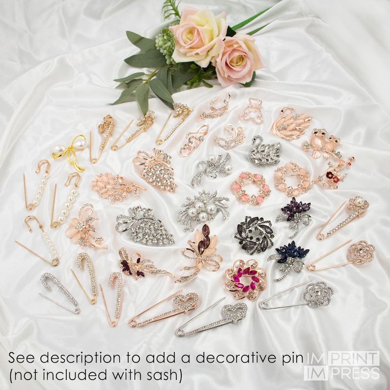 May include: Assortment of decorative pins with rhinestones and pearls. The pins are in various shapes, including flowers, swans, and leaves. The pins are displayed on a white satin fabric background. See description to add a decorative pin (not included with sash). IM PRINT IM PRESS