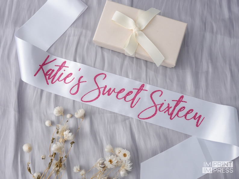 May include: White satin sash with pink glitter lettering that reads "Katie's Sweet Sixteen".