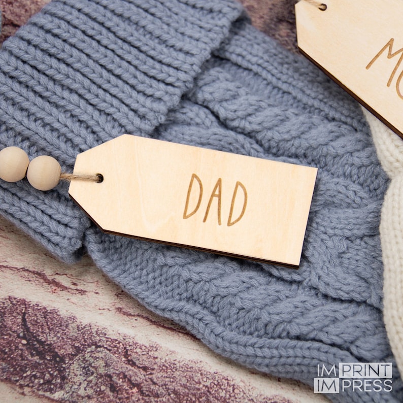 K&ouml;nnte beinhalten: Ein h&ouml;lzerner Geschenkanh&auml;nger mit dem eingravierten Wort "DAD". Der Anh&auml;nger ist an einer grauen Strickm&uuml;tze mit Holzperlen befestigt.