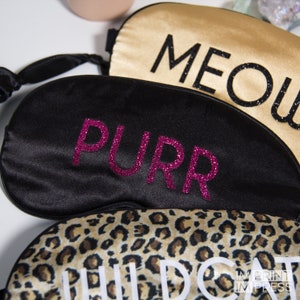 Op de afbeelding: Drie zwarte satijnen slaapmaskers met verschillende tekst. Het bovenste masker zegt "MEOW" in zwart glitter. Het middelste masker zegt "PURR" in roze glitter. Het onderste masker heeft een luipaardprint en zegt "WILD CAT" in witte tekst.