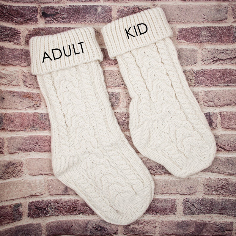 K&ouml;nnte beinhalten: Zwei wei&szlig;e Strickstr&uuml;mpfe mit Zopfmuster. Der Strumpf links hat das Wort "ADULT" auf der Manschette gestickt. Der Strumpf rechts hat das Wort "KID" auf der Manschette gestickt.