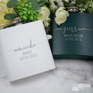 Puede incluir: Dos  flascos de cadera de acero inoxidable personalizados con un acabado blanco y verde oscuro. El frasco blanco tiene el nombre "amanda" y el texto "BRIDE 07.02.2022". El frasco verde oscuro tiene el nombre "sierra" y el texto "BRIDESMAID 07.02.2022".