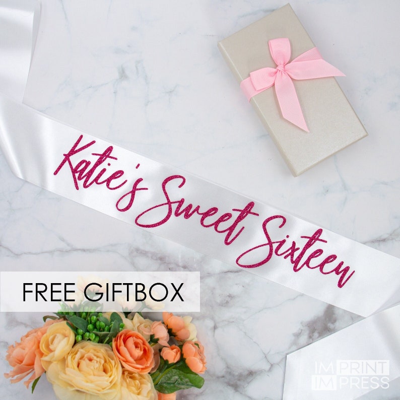 May include: White satin sash with pink glitter lettering that reads "Katie's Sweet Sixteen". The sash is displayed on a white marble surface with a small gift box and flowers.