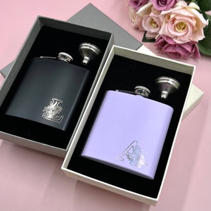 May include: Two personalized hip flasks in gift boxes. One flask is black with a silver 'L' and floral design. The other flask is lavender with a silver 'A' and floral design.