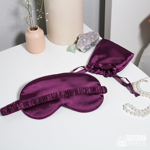 May include: A purple satin sleep mask with an elastic band. The mask is lying on a white surface with a small purple satin bag and a pearl necklace.