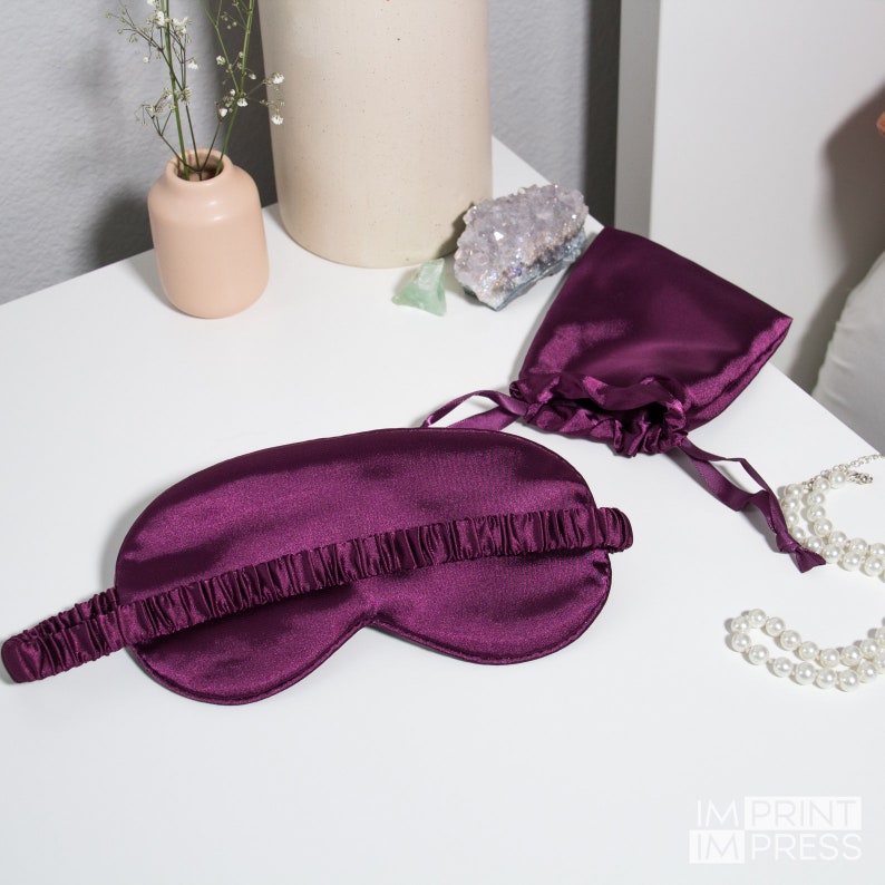May include: A purple satin sleep mask with an elastic band. The mask is lying on a white surface with a small purple satin bag and a pearl necklace.