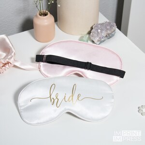 May include: Two satin sleep masks, one pink and one white. The white mask has the word "bride" written in gold script.