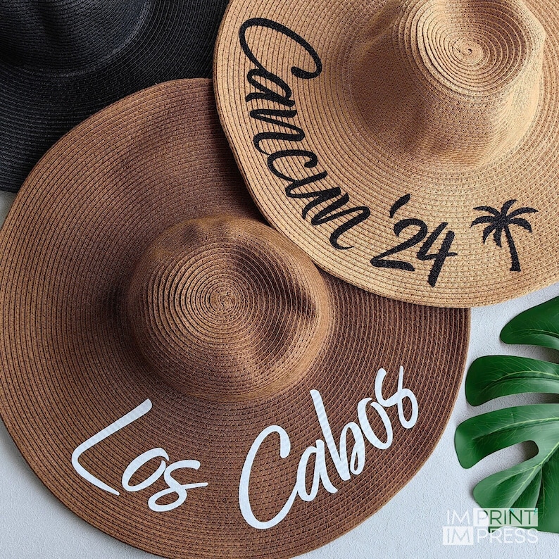 May include: Two brown straw sun hats with white and black text. The hat on the left says 'Los Cabos' in white text. The hat on the right says 'Cancun '24' in black text with a palm tree icon.