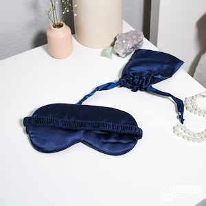 May include: A navy blue satin sleep mask with an elastic strap. The mask is folded and placed on a white surface with a small pouch next to it.
