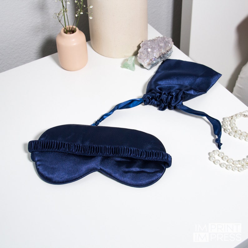 May include: A navy blue satin sleep mask with an elastic strap. The mask is folded and placed on a white surface with a small pouch next to it.