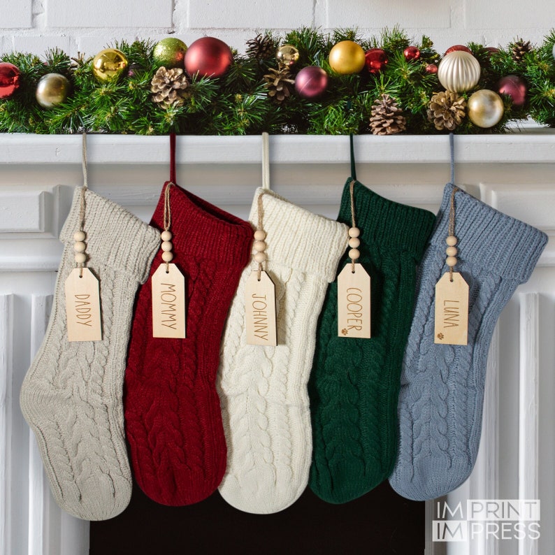 K&ouml;nnte beinhalten: F&uuml;nf personalisierte Weihnachtsstr&uuml;mpfe h&auml;ngen an einem Kaminsims. Die Str&uuml;mpfe sind aus gestricktem Stoff in verschiedenen Farben, darunter Beige, Rot, Wei&szlig;, Gr&uuml;n und Grau. Jeder Strumpf hat ein Holzschild mit einem Namen darauf: Papa, Mama, Johnny, Cooper und Luna.