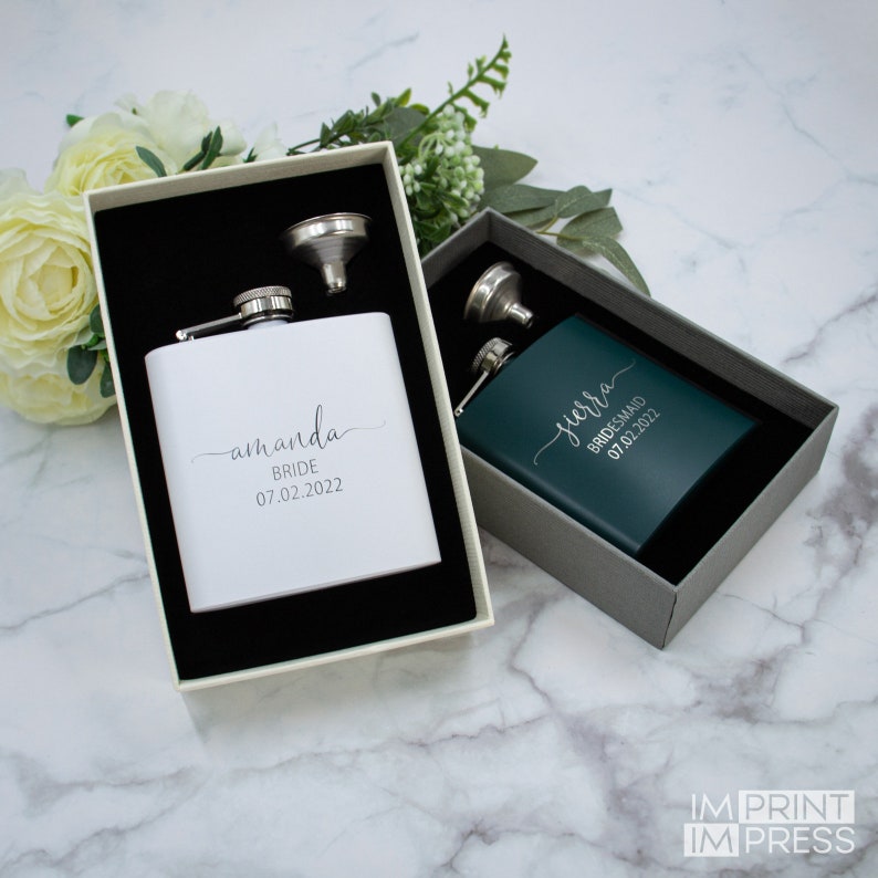 Puede incluir: Dos matraces de acero inoxidable personalizados con embudos en cajas de regalo. El matraz de la izquierda es blanco con el texto "amanda BRIDE 07.02.2022". El matraz de la derecha es verde con el texto "jerra BRIDESMAID 07.02.2022".