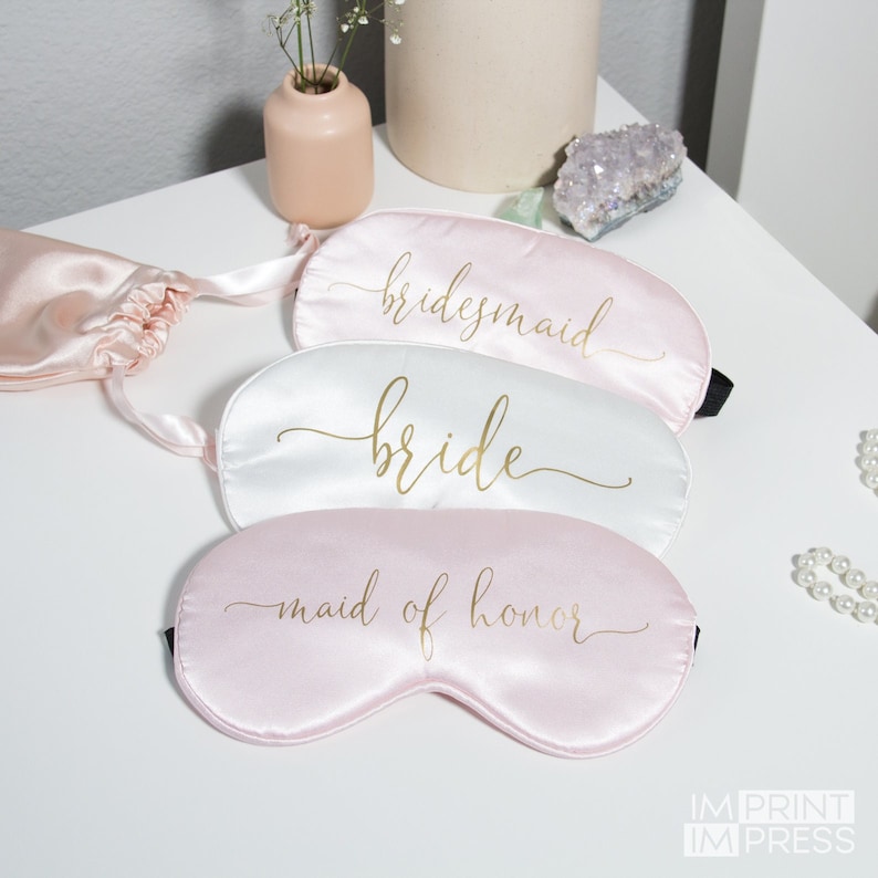 May include: Three satin sleep masks with gold lettering. The top mask is pink and says "bridesmaid". The middle mask is white and says "bride". The bottom mask is pink and says "maid of honor".