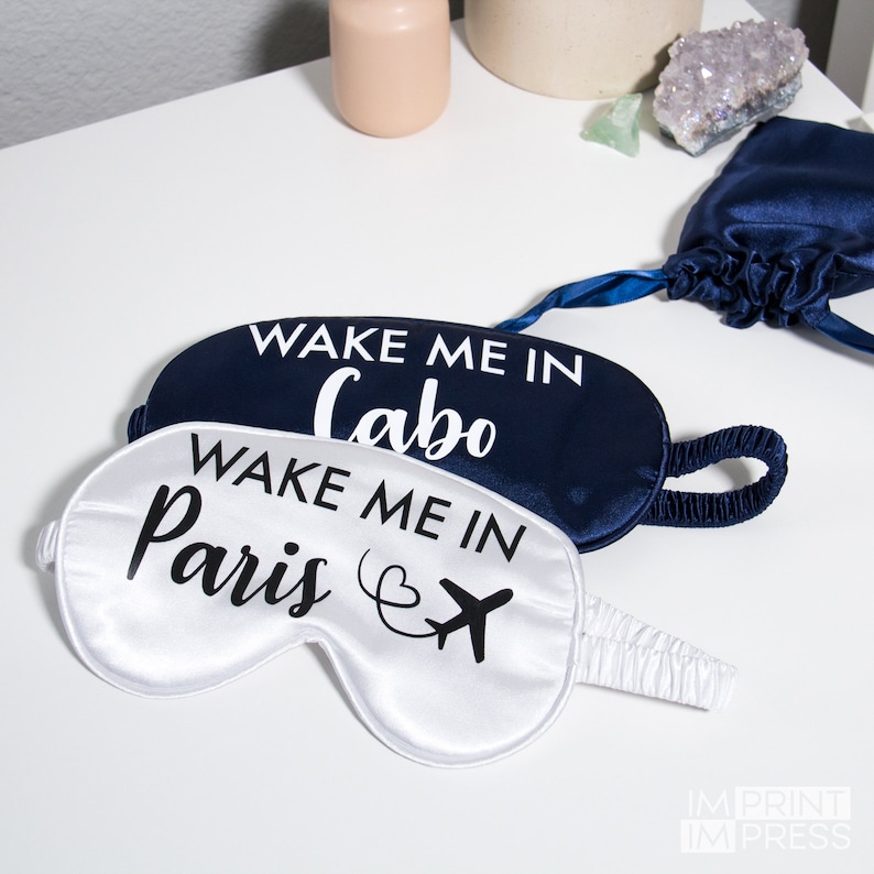 May include: Two satin sleep masks with the text "Wake me in Cabo" and "Wake me in Paris" printed on them. The mask with the text "Wake me in Cabo" is navy blue and the mask with the text "Wake me in Paris" is white.