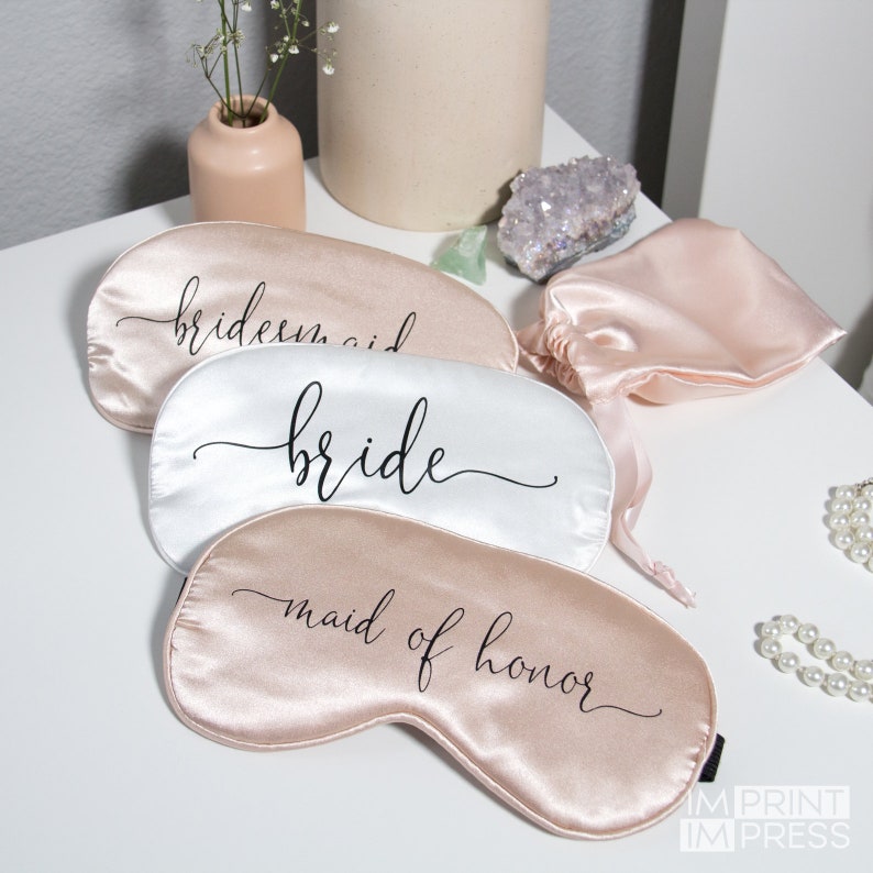 May include: Three satin sleep masks with the words "bride", "bridesmaid", and "maid of honor" printed in black script. The masks are in a light pink and white color scheme.