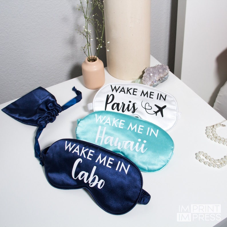 May include: Three satin sleep masks with the text "Wake Me In" followed by a city name. The masks are white, light blue, and dark blue. The text is in black.