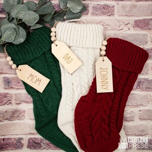 K&ouml;nnte beinhalten: Drei gestrickte Weihnachtsstr&uuml;mpfe mit Holzetiketten. Die Str&uuml;mpfe sind gr&uuml;n, wei&szlig; und rot. Die Etiketten sind mit "Mama", "Papa" und "Johnny" beschriftet.