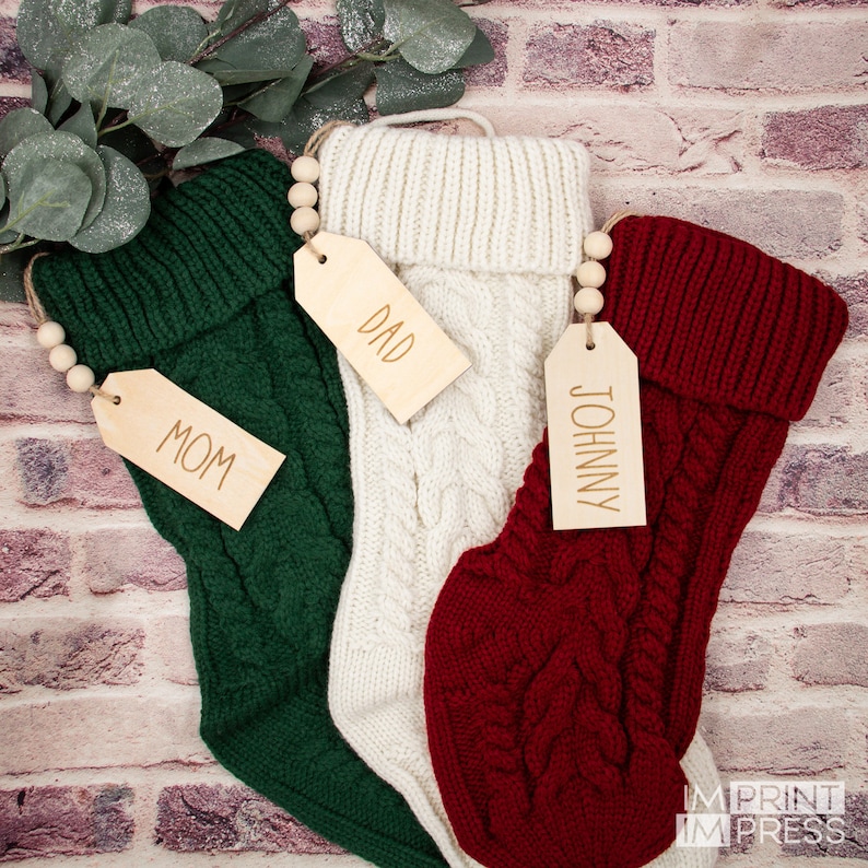 K&ouml;nnte beinhalten: Drei gestrickte Weihnachtsstr&uuml;mpfe mit Holzetiketten. Die Str&uuml;mpfe sind gr&uuml;n, wei&szlig; und rot. Die Etiketten sind mit "Mama", "Papa" und "Johnny" beschriftet.