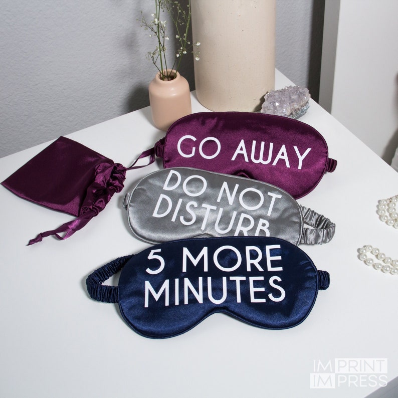 May include: Three satin sleep masks with different phrases printed on them. The top mask is purple and says "Go Away". The middle mask is silver and says "Do Not Disturb". The bottom mask is navy blue and says "5 More Minutes".