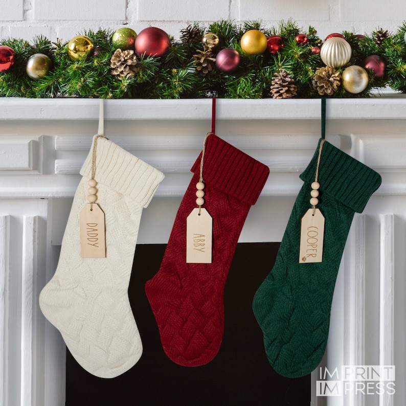 May include: Three knitted Christmas stockings with personalized wooden tags hanging from a white fireplace mantel. The stockings are cream, red, and green. The tags read "Daddy", "Abby", and "Cooper".