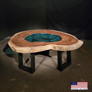 May include: A large, natural wood coffee table with a unique, irregular shape. The table top features a large, teal-colored resin inlay. The table is supported by four black metal legs.