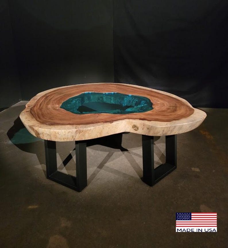 May include: A large, natural wood coffee table with a unique, irregular shape. The table top features a large, teal-colored resin inlay. The table is supported by four black metal legs.