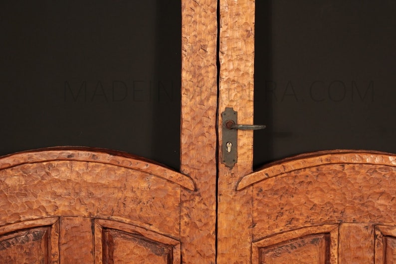 Puede incluir: Primer plano de una puerta doble de madera con un dise&ntilde;o tallado. La puerta tiene una manija de puerta de metal y una cerradura. La puerta est&aacute; hecha de madera marr&oacute;n claro con un acabado r&uacute;stico.