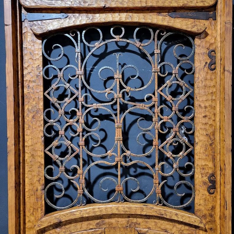 May include: A wooden door with a decorative iron grate. The grate is made of intricate swirls and loops, and it is set into the door in a rounded arch. The wood is a warm brown colour, and the iron is a dark grey.