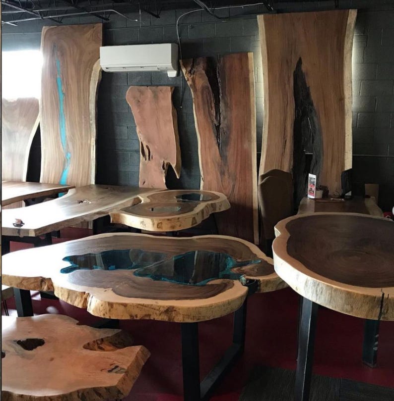 May include: A collection of large wooden slabs with live edge details, some with epoxy resin inlays. The slabs are arranged in a workshop setting, showcasing their natural beauty and unique character.