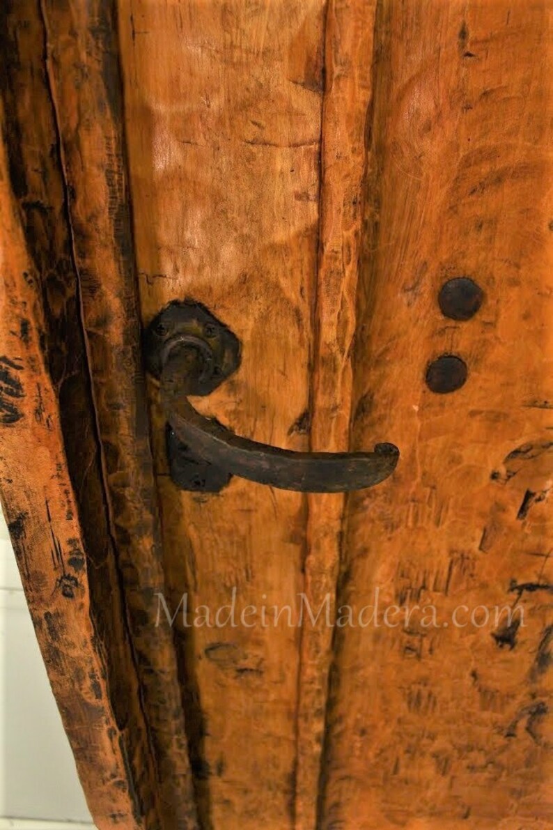 Puede incluir: Primer plano de una puerta de madera con un acabado marr&oacute;n oscuro envejecido. La puerta tiene un tirador de metal negro y dos bisagras de metal negro. El texto "MadeinMadera.com" es visible en la puerta.