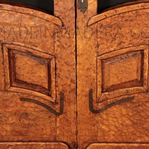 Puede incluir: Primer plano de un par de puertas de madera con intrincados tallados y detalles met&aacute;licos. Las puertas son de un color marr&oacute;n intenso y tienen un aspecto r&uacute;stico y antiguo. El texto "MADE IN" est&aacute; tallado en la parte superior de las puertas.