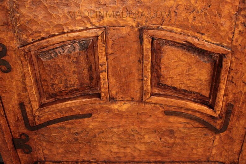 Puede incluir: Primer plano de una puerta de armario de madera con dos paneles y bisagras de hierro decorativas. La madera es de un color marr&oacute;n intenso y tiene una superficie rugosa y texturizada.