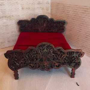 Puede incluir: Una cama en miniatura con cabecero y piecero ornamentados de color marrón oscuro. El marco de la cama tiene un diseño tallado detallado con toques de verde. La cama está cubierta con una manta de color rojo intenso. Mueble de casa de muñecas.