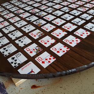 May include: A wooden circular game board with a brown finish. The board is covered with playing cards, each with a different number and suit. The cards are arranged in a grid pattern. The board is on a wooden surface.