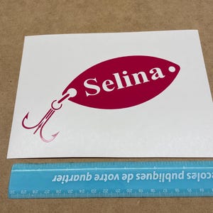 May include: A pink and white vinyl decal of a fishing lure with the name "Selina" written on it.
