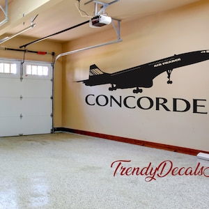 May include: Black and white silhouette of a Concorde airplane with the text "AIR FRANCE" and "CONCORDE" on a beige wall in a garage. The text "Trendy Decals Art" is on the floor.