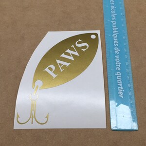 May include: Gold-colored fish-shaped sticker with the word "PAWS" in white lettering. The sticker has a hook and two treble hooks.