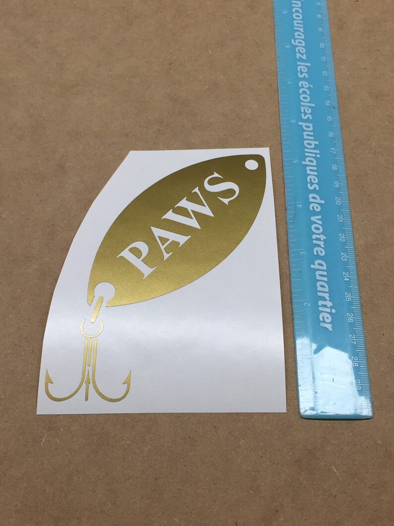 May include: Gold-colored fish-shaped sticker with the word "PAWS" in white lettering. The sticker has a hook and two treble hooks.